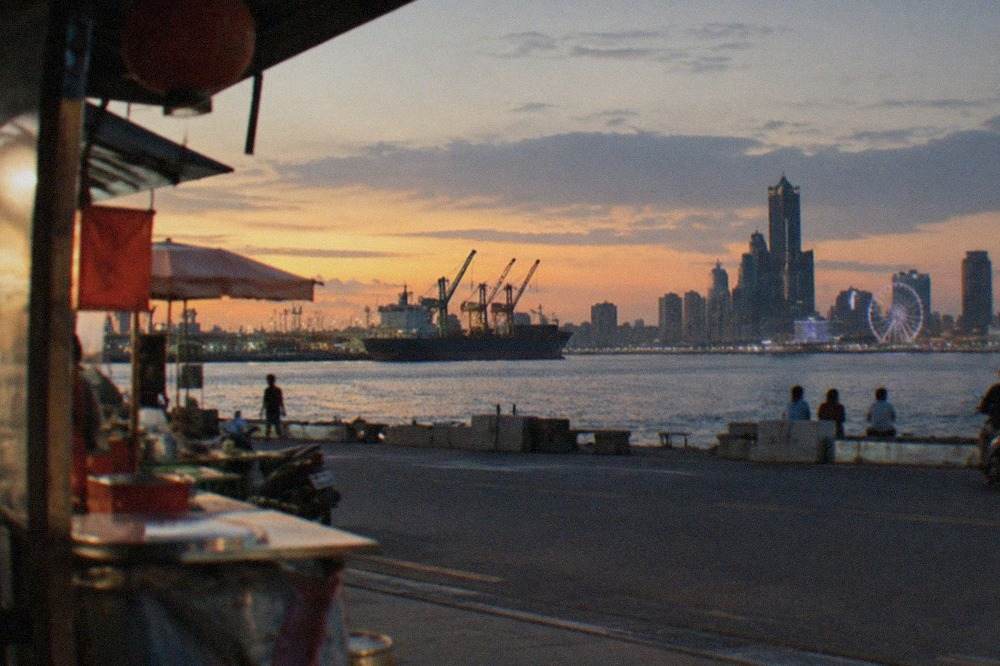 A wide harbor skyline at dusk with cargo cranes, a ferris wheel and illuminated buildings along the water