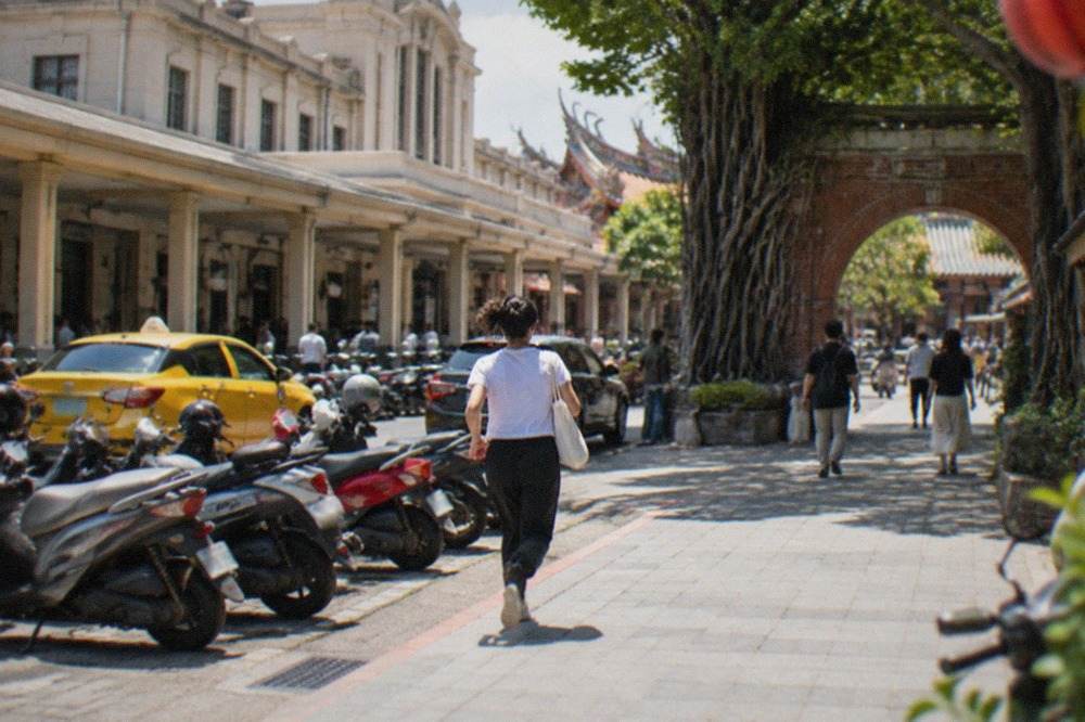 A historic brick archway with banyan roots and visitors walking through a sunlit old Taiwanese courtyard