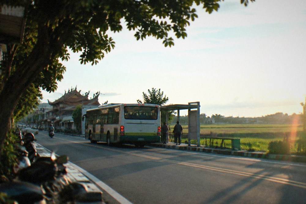 A city bus stopped at a roadside stop beside wide flat farmland under a blue sky