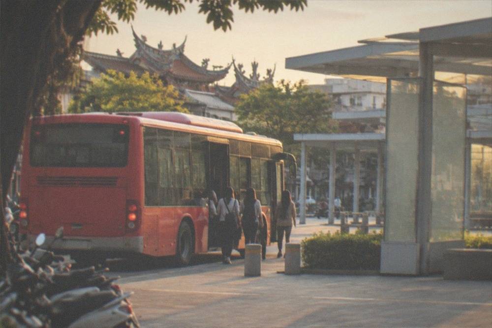A red city bus at a stop with passengers boarding beside a modern station plaza