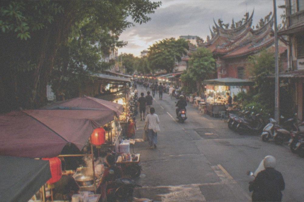An evening street lined with food stalls, glowing signs and customers browsing under lights