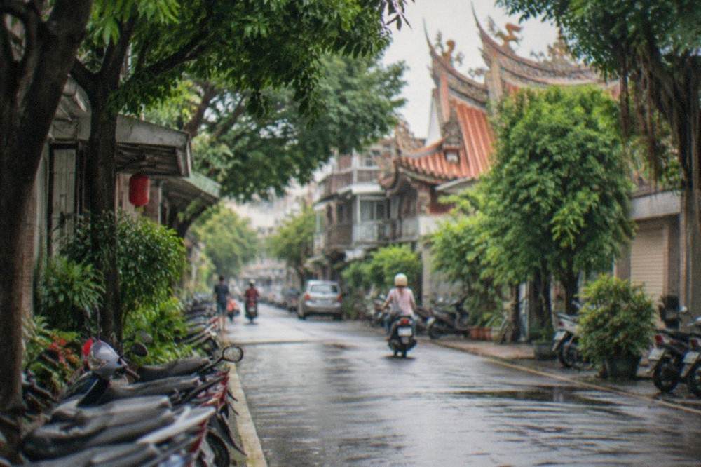 A quiet residential street with low buildings, scooters parked at the curb and green trees shading the sidewalk