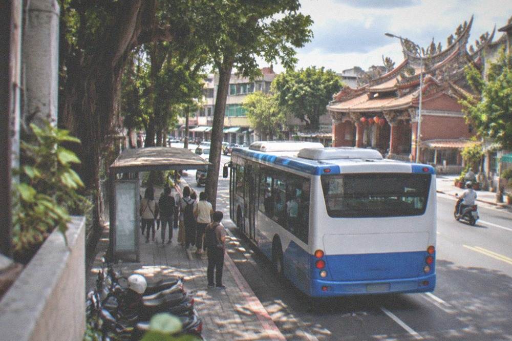 A blue-and-white city bus at a roadside stop with passengers waiting in the shade