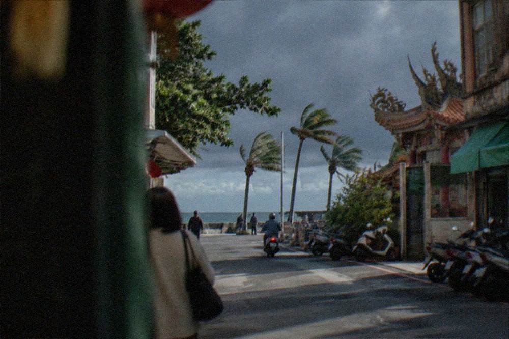 Dark storm clouds gathering over a flat coastal horizon with wind-bent palm trees along a seafront path