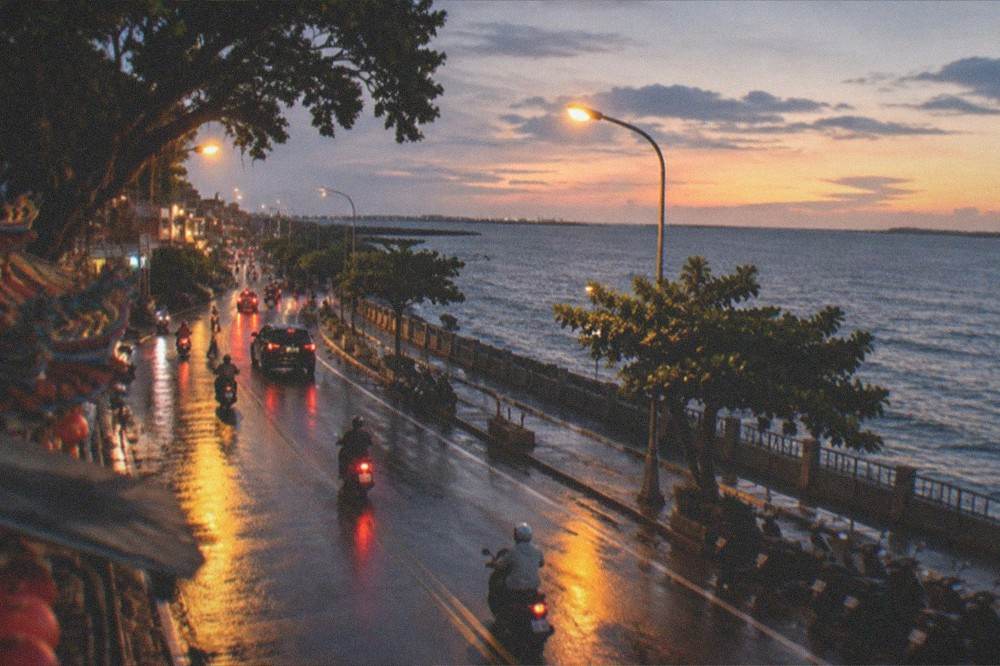 A seafront road at dusk with streetlights reflecting on wet pavement and a calm dark sea beyond