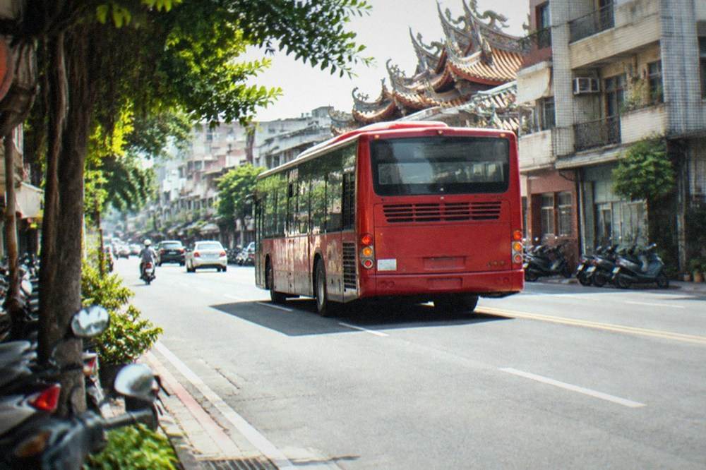 A red city bus pulling away from a stop on a wide Taiwanese road with apartment buildings behind it