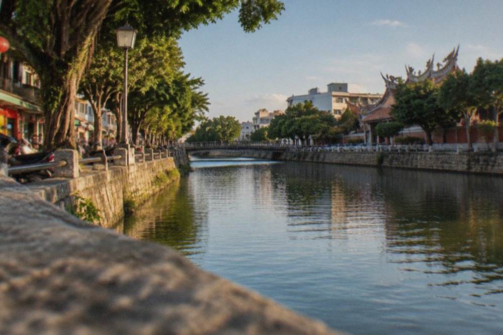 A wide canal in Anping, Tainan, with a stone embankment, trees along the bank and buildings in the distance under a blue sky