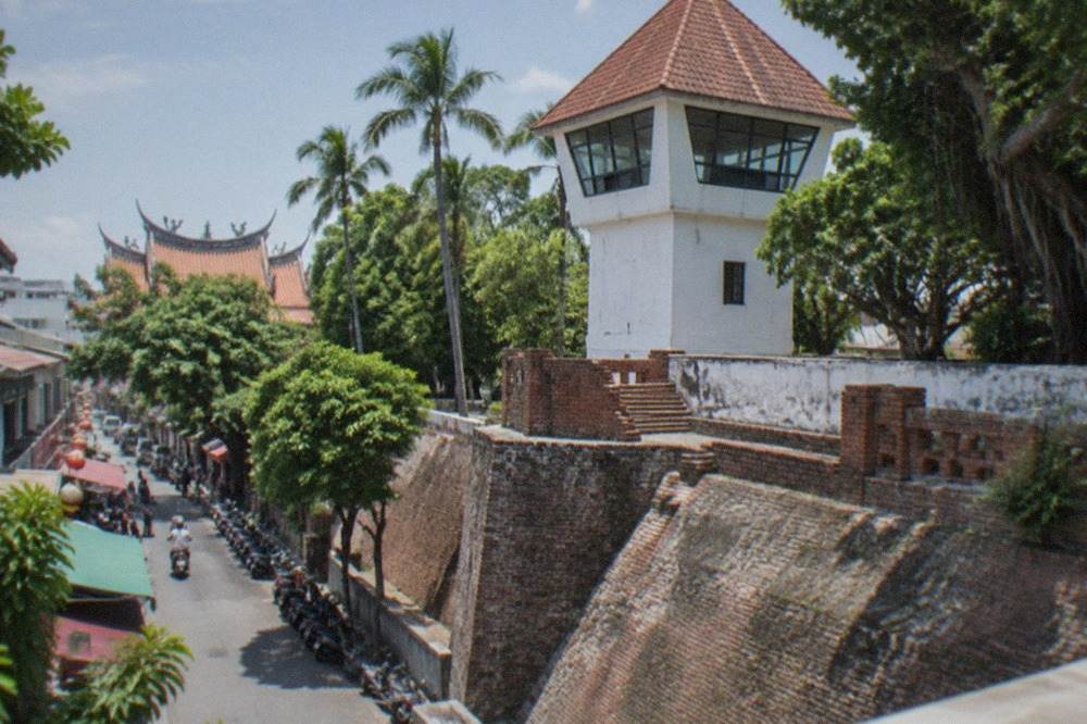 A white-walled fort lookout tower with a pointed red-tiled roof rising above old brick ramparts and palm trees
