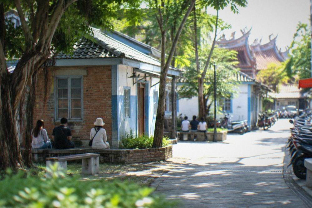 Visitors resting under trees beside a low brick building in a park during warm weather