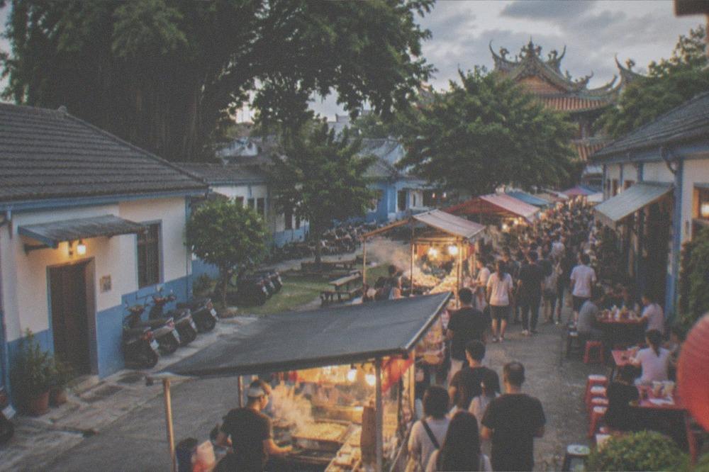 A crowded night market aisle lit by stall lights, with food vendors cooking and people eating at small tables
