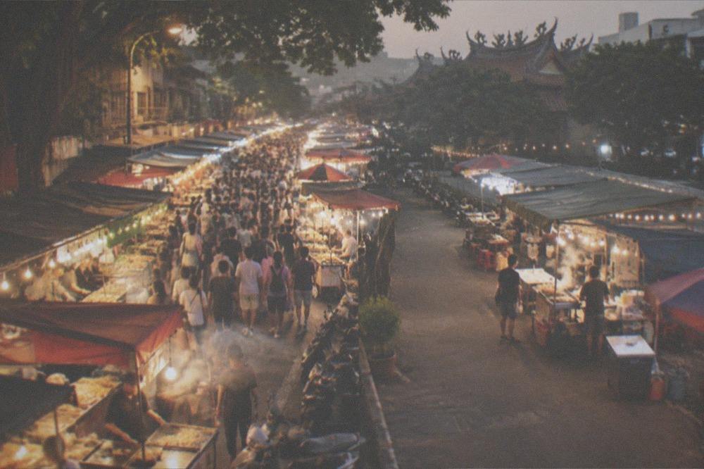 Two night markets side by side in the frame, one crowded and one quieter, under strings of white bulbs