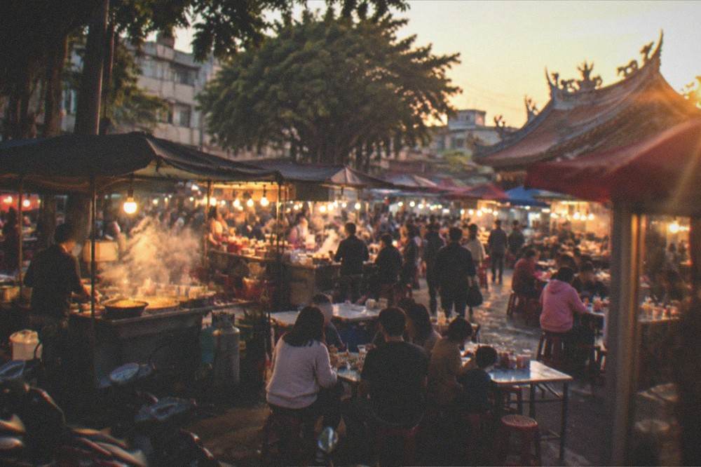 Families and friends sharing food at plastic tables under market lights, with steam rising from stalls behind them