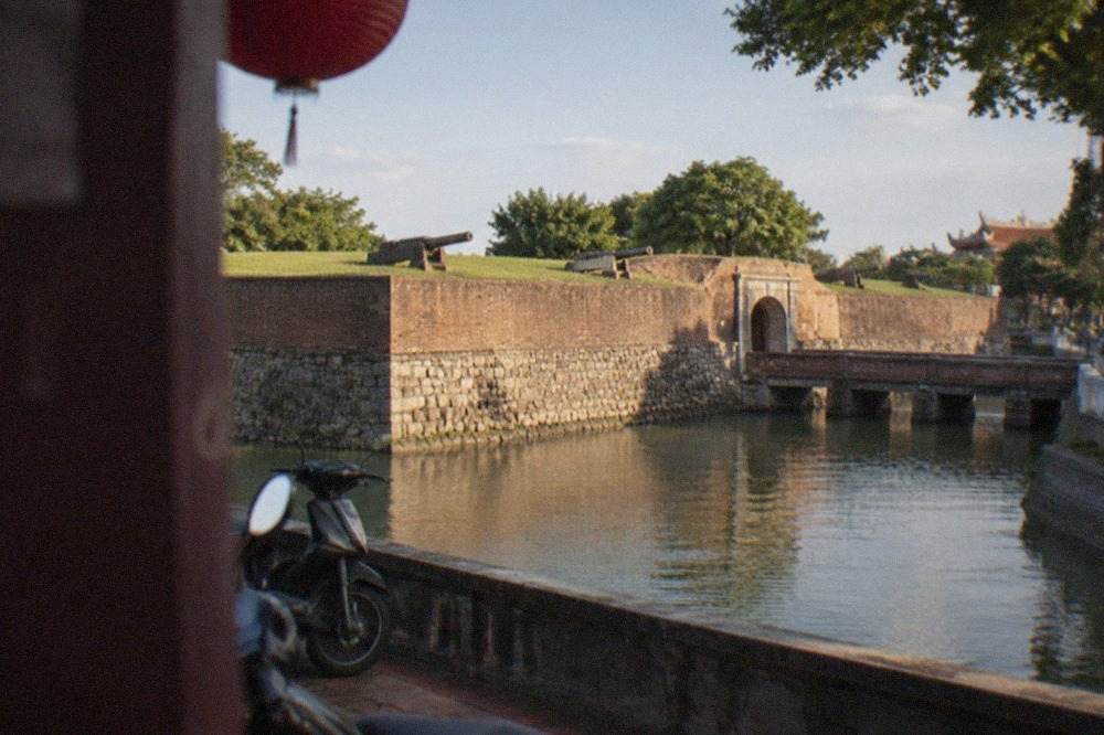 A low stone fort with grassy ramparts and an arched entrance set beside a wide moat under a bright sky