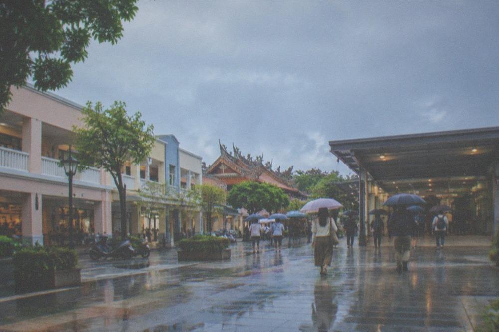 Rain falling on a station plaza with travelers moving toward a covered entrance