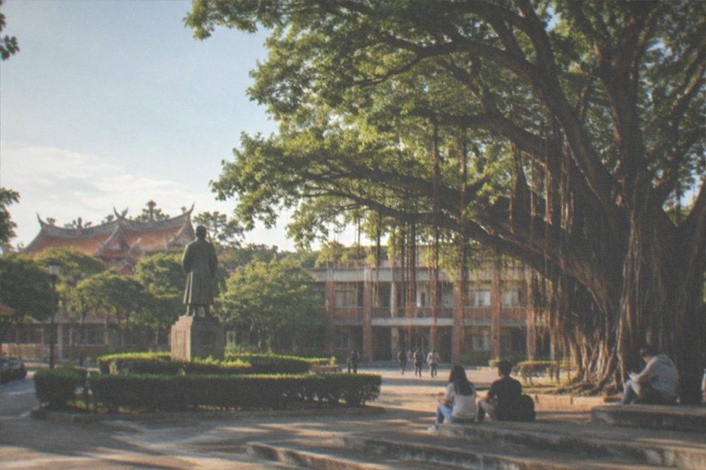 A quiet campus courtyard with a statue, trimmed hedges and students sitting on steps