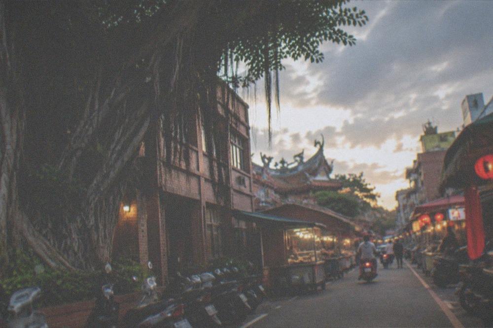 A narrow food street at dusk with lit stall signs and people queuing at counters