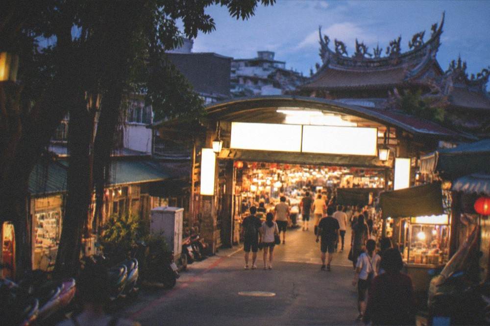A covered market entrance at night, warm light spilling from inside and people arriving in ones and twos, the illuminated boards above blown out and unreadable
