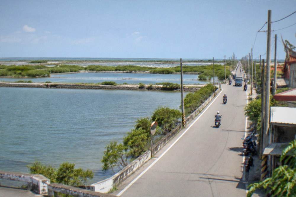 A flat coastal road running between fish ponds toward distant wetlands under a blue sky