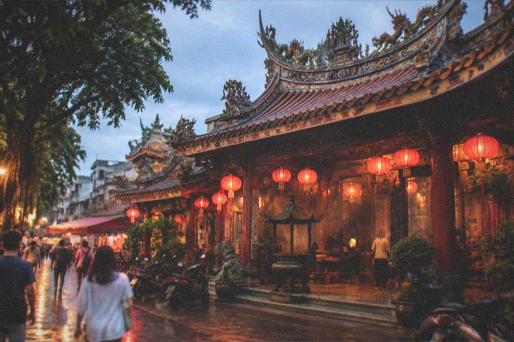 Wide view of a Tainan temple facade at dusk with lit lanterns and people walking past in the warm street light