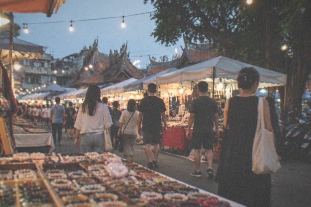 Rows of market stalls under string lights with shoppers walking between them