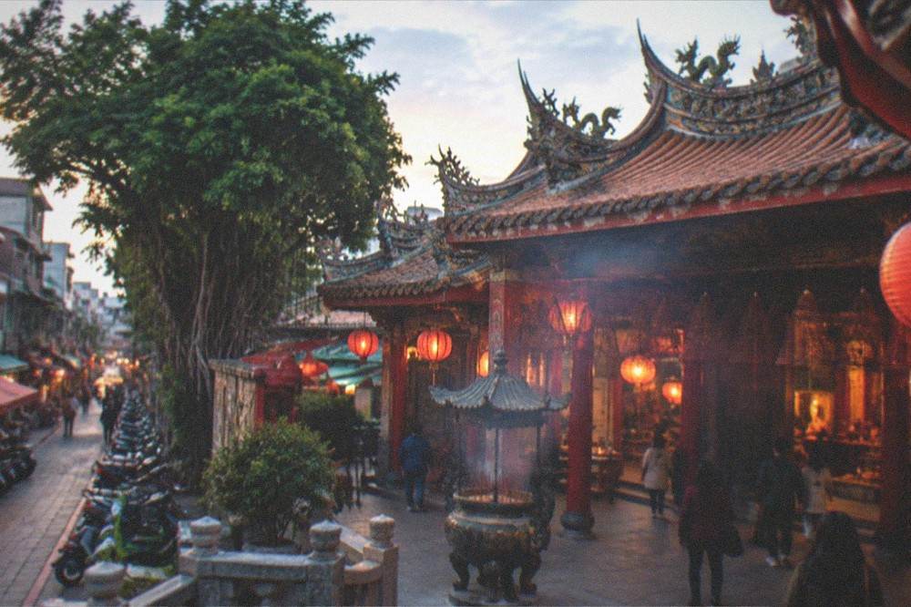 A lit Taiwanese temple facade at dusk with warm lanterns and visitors on the forecourt