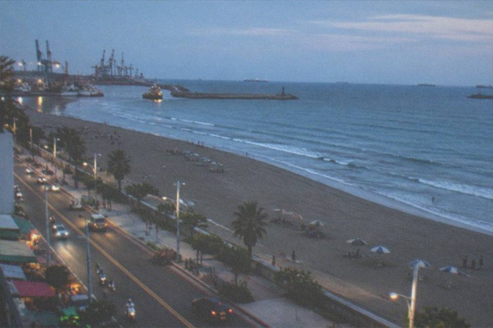 A wide sandy beach with gentle waves, umbrellas and a low seawall along a coastal road