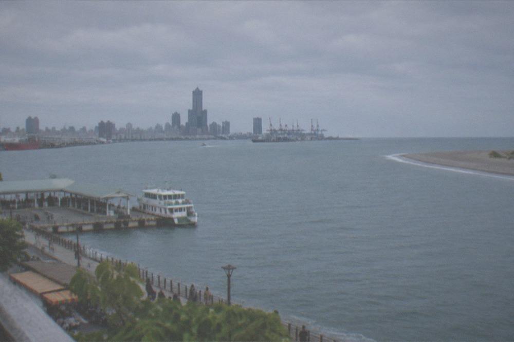 A small ferry at a harbor pier with a city skyline of towers and cranes across the water