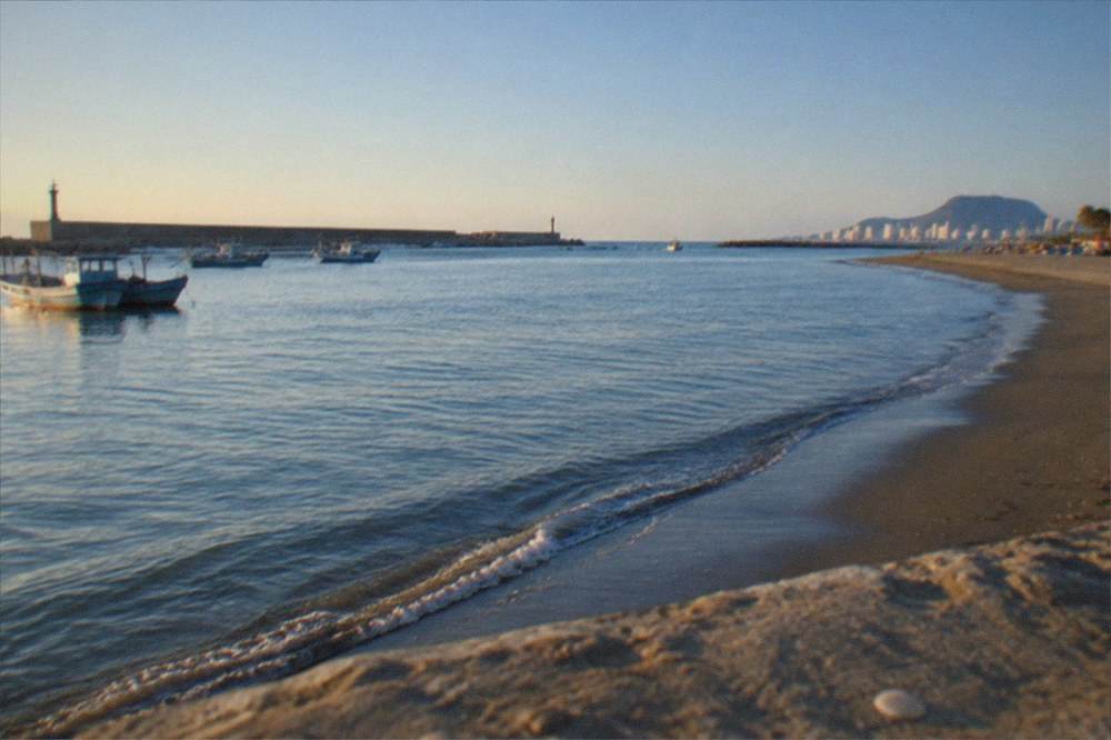 A coastal view under a clear sky with calm water, a long breakwater and small boats near shore