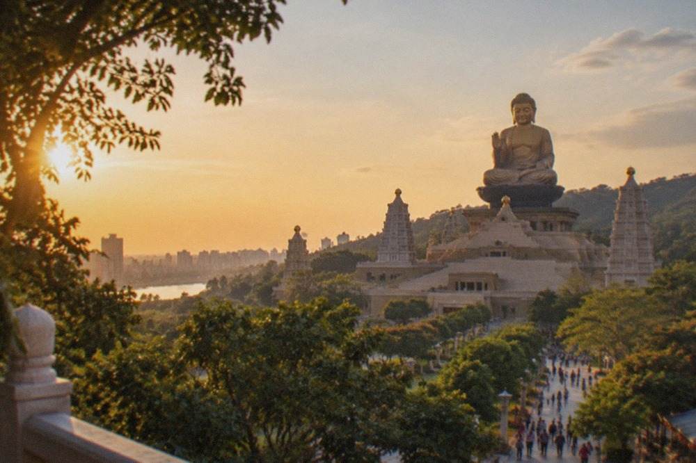 A giant seated Buddha statue in front of a large monastery building surrounded by trees