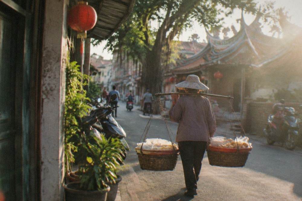 A street vendor using a shoulder pole to carry two baskets of food through a narrow lane