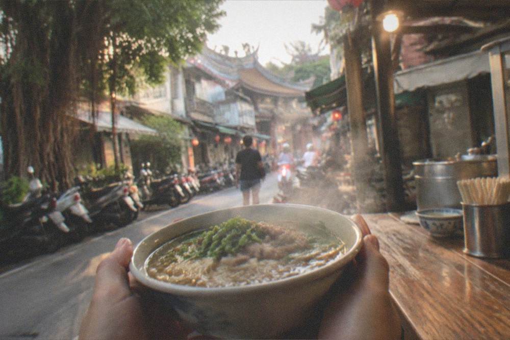 A steaming small noodle bowl held in two hands above a wooden street stall counter at night