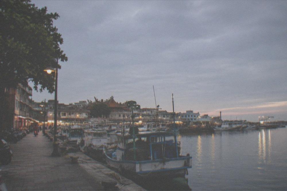 A harbor front at dusk with fishing boats and a lit promenade