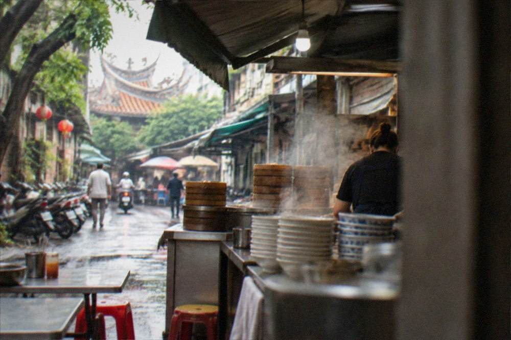 A food stall counter with steaming bowls stacked at the pass and a cook working behind it, any price board out of frame