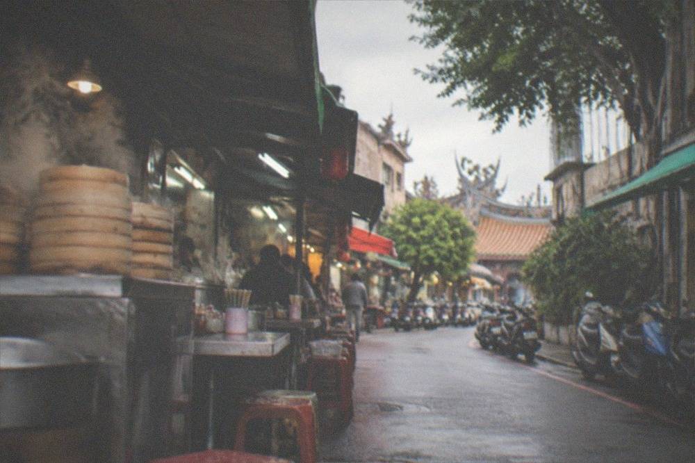 A narrow Tainan street in the morning with small food stalls and scooters parked along the curb