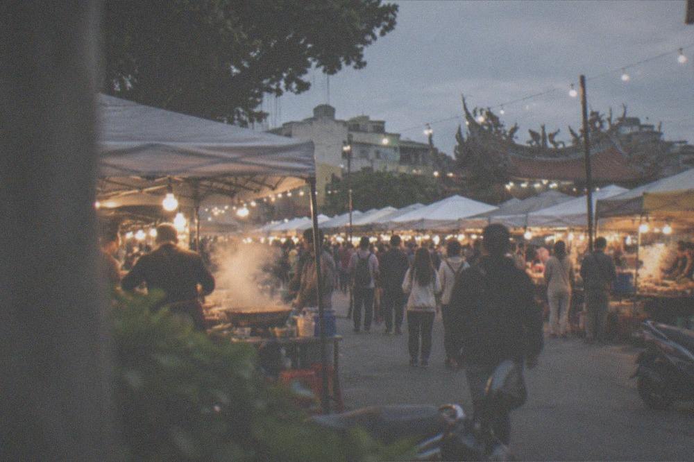Crowded night market stalls lit by warm bulbs with steam rising from food carts