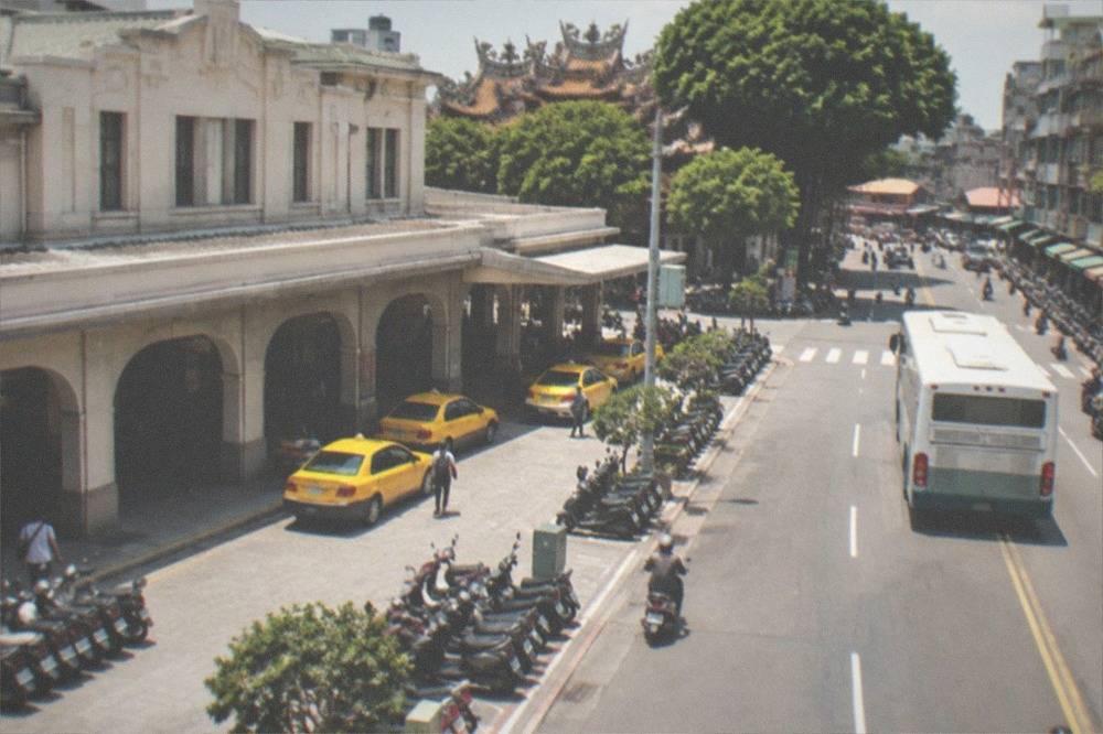 A city bus pulling away from a stop on a broad street lined with shopfronts and scooters
