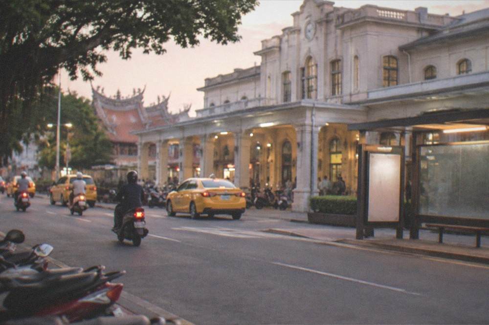 A bus stop at dusk with a lit timetable sign and a street of glowing shopfronts behind it