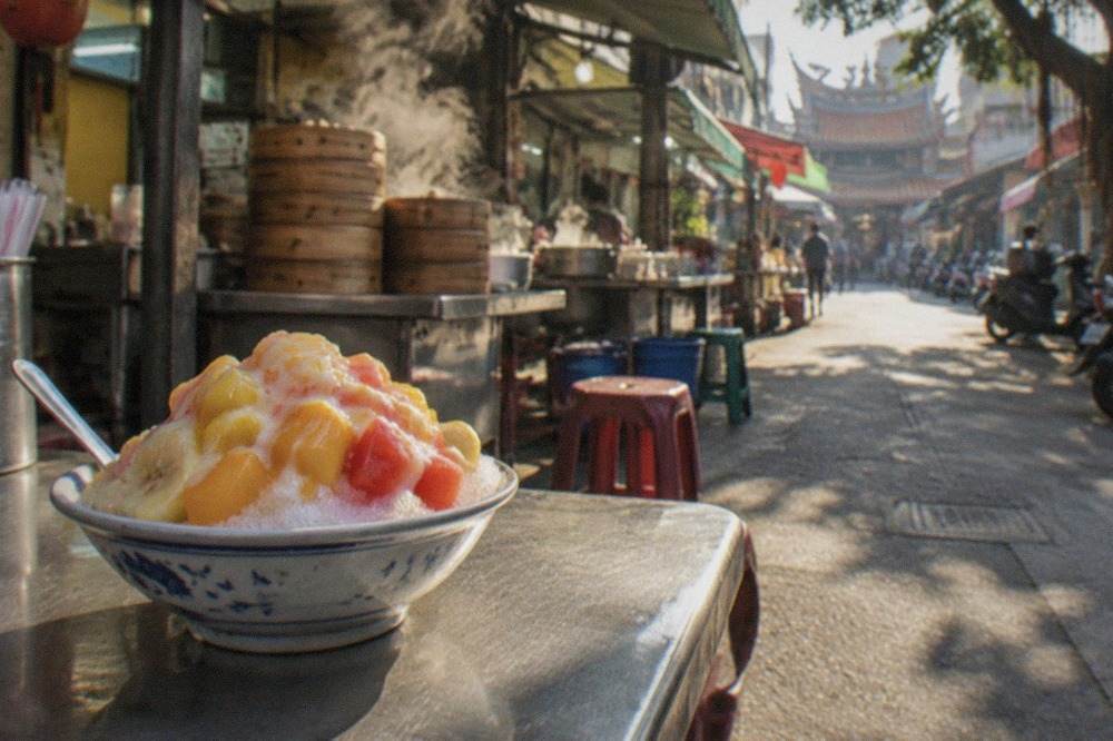 A bowl of Taiwanese shaved ice topped with fruit and condensed milk on a market table