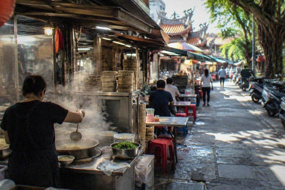 A cook ladling broth over noodles at a small street food stall in Tainan