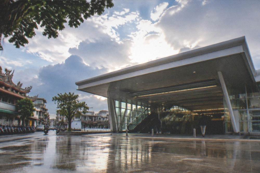 A station entrance plaza under a clearing sky after rain, with wet paving reflecting the building