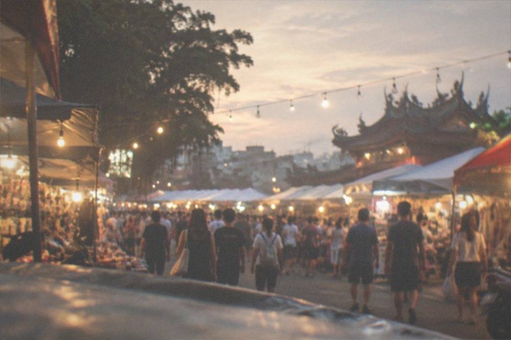 A wide view of a night market lot at dusk, with rows of lit stalls and a dense crowd walking between them