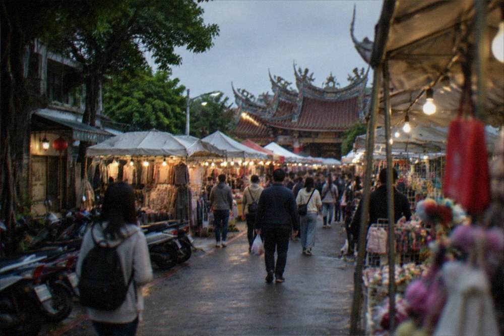 Night market stalls under a covered roof structure, with shoppers walking in a dry indoor arcade lit by stall lights