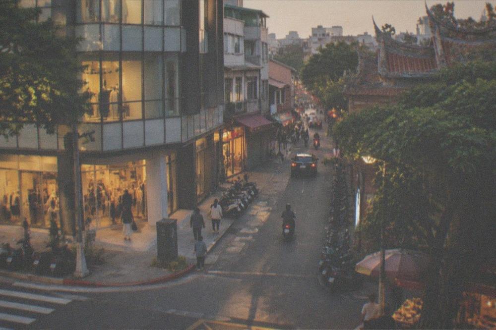 A Tainan shopping street at dusk, shutters half down and warm light spilling from a doorway, shopfront boards unlit and indistinct
