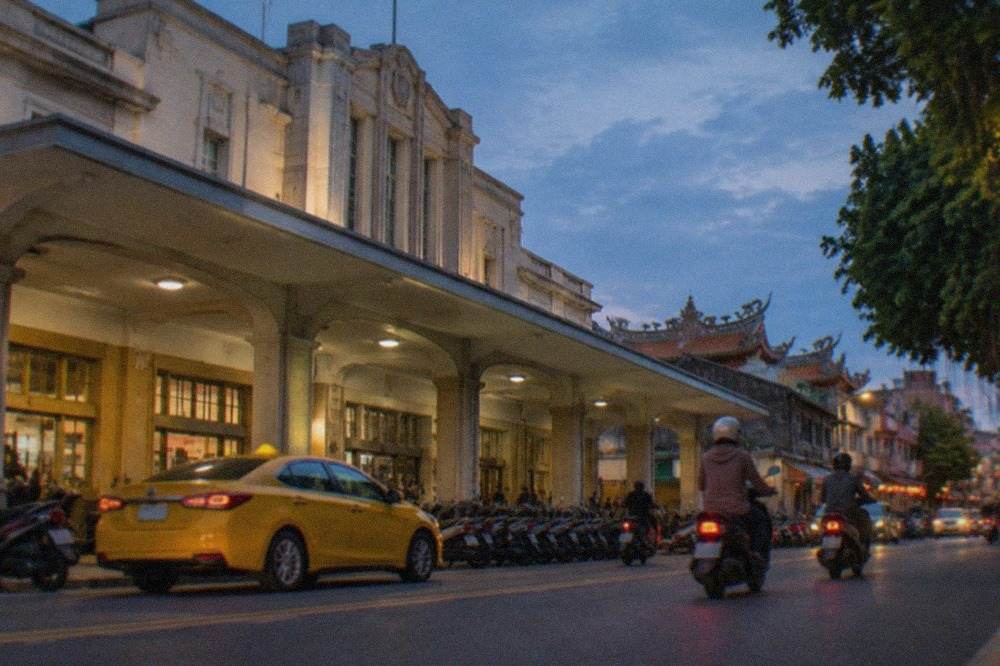 A yellow taxi waiting on a street in central Tainan with scooters passing by