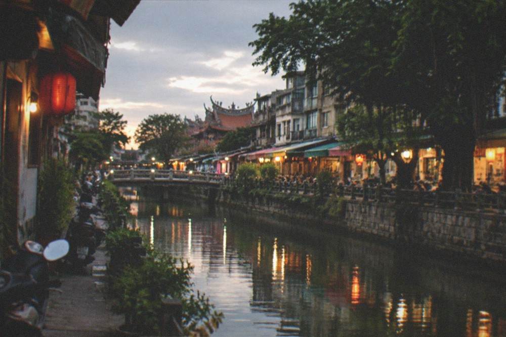 A canal-side lane in Tainan at dusk with warm shop lights reflecting on the water