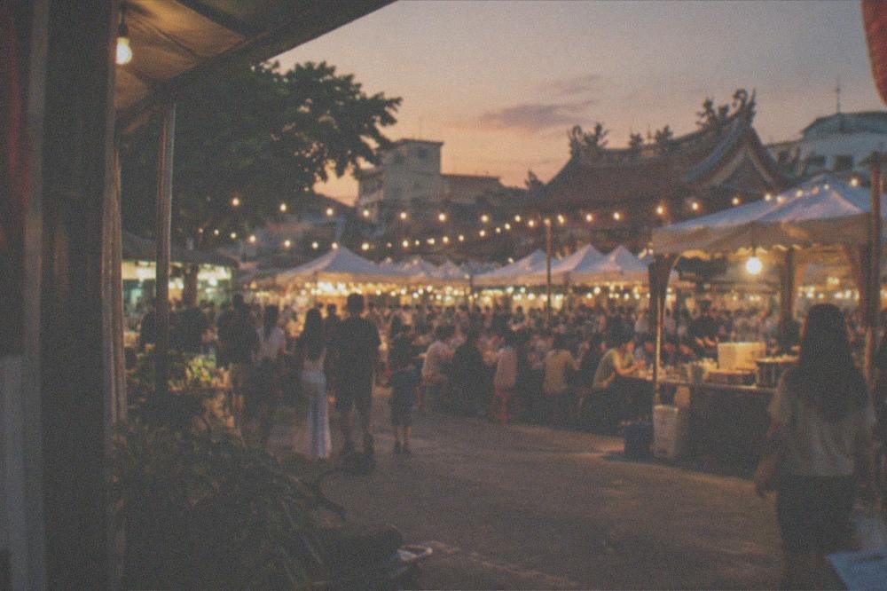 Families eating at crowded food stalls under string lights at an outdoor night market