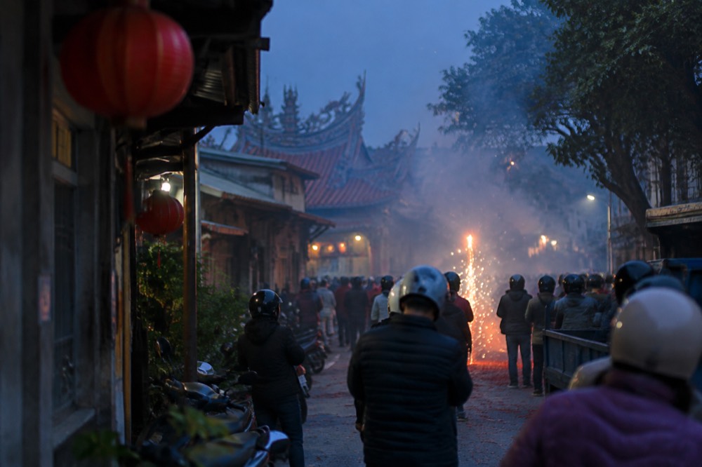 A crowd of people in full-face helmets and padded jackets standing in a smoky street lit by fireworks
